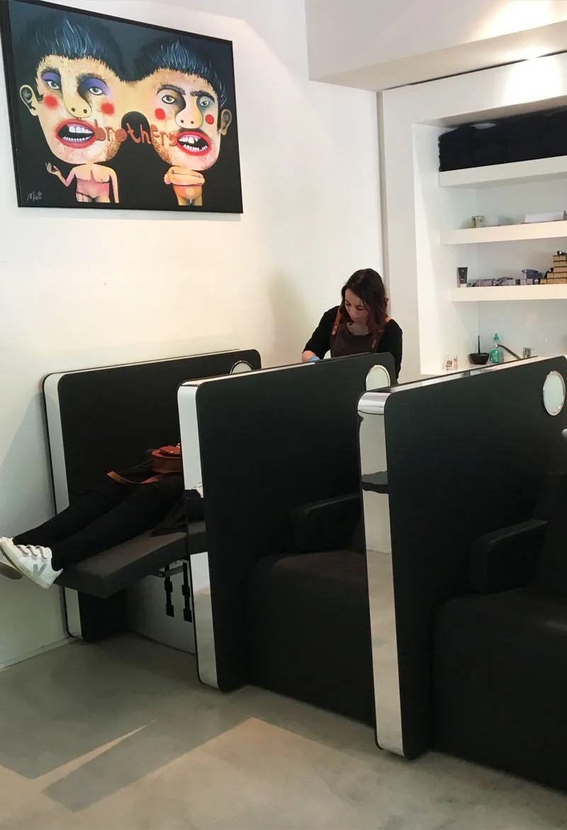 Les bacs de lavage du salon, alignés sous une grande toile figurative, une coiffeuse préparant le poste au fond.