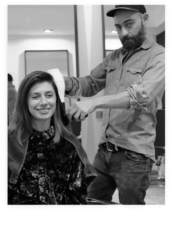 Un coiffeur du salon réalise un brushing sur une cliente souriante, photographie en noir et blanc.