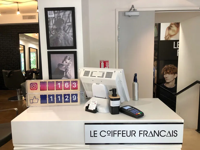 Le comptoir d’accueil du salon, portant le nom Le Coiffeur Français, avec le terminal de paiement et l’entrée du salon derrière.