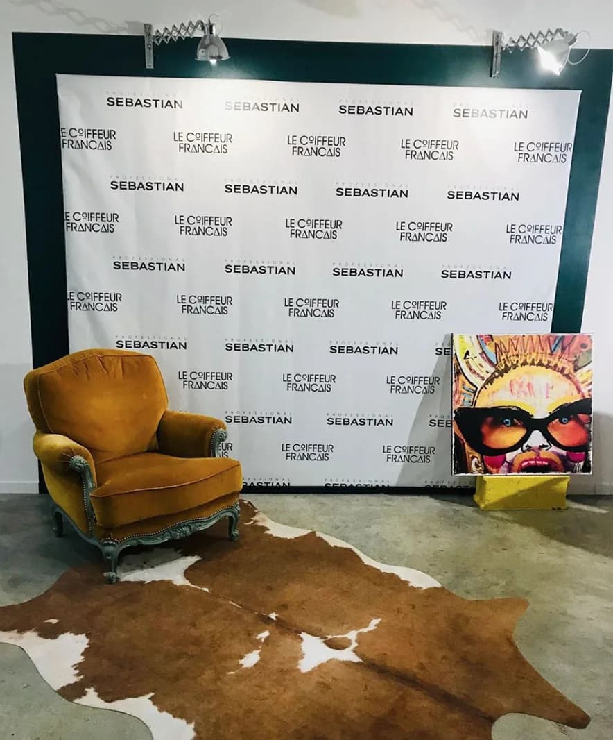 Un fauteuil de velours ocre sur un tapis en peau, devant un mur portant les noms Le Coiffeur Français et Sebastian, à côté d’une toile pop.