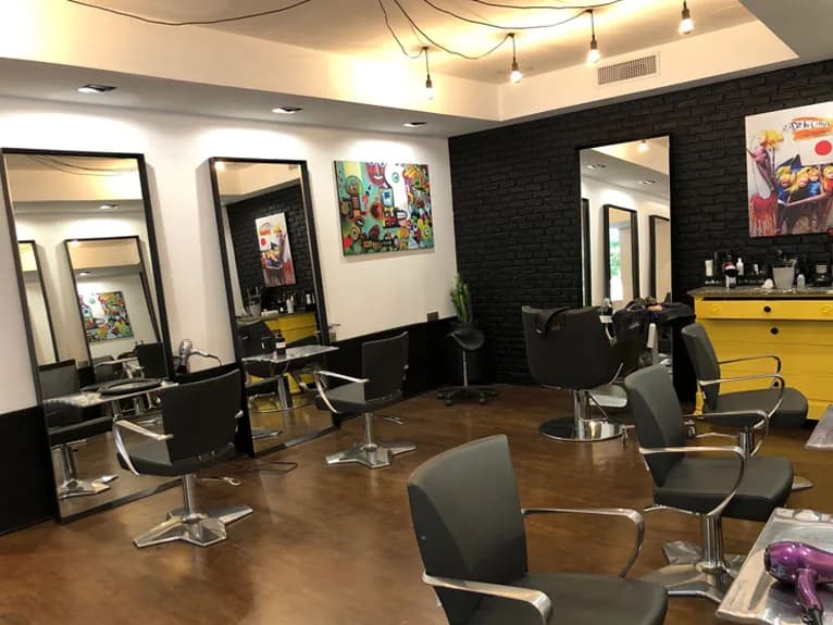 La salle de coiffage : miroirs à cadre noir, fauteuils gris, mur de brique peint en noir, toiles colorées et meuble jaune.